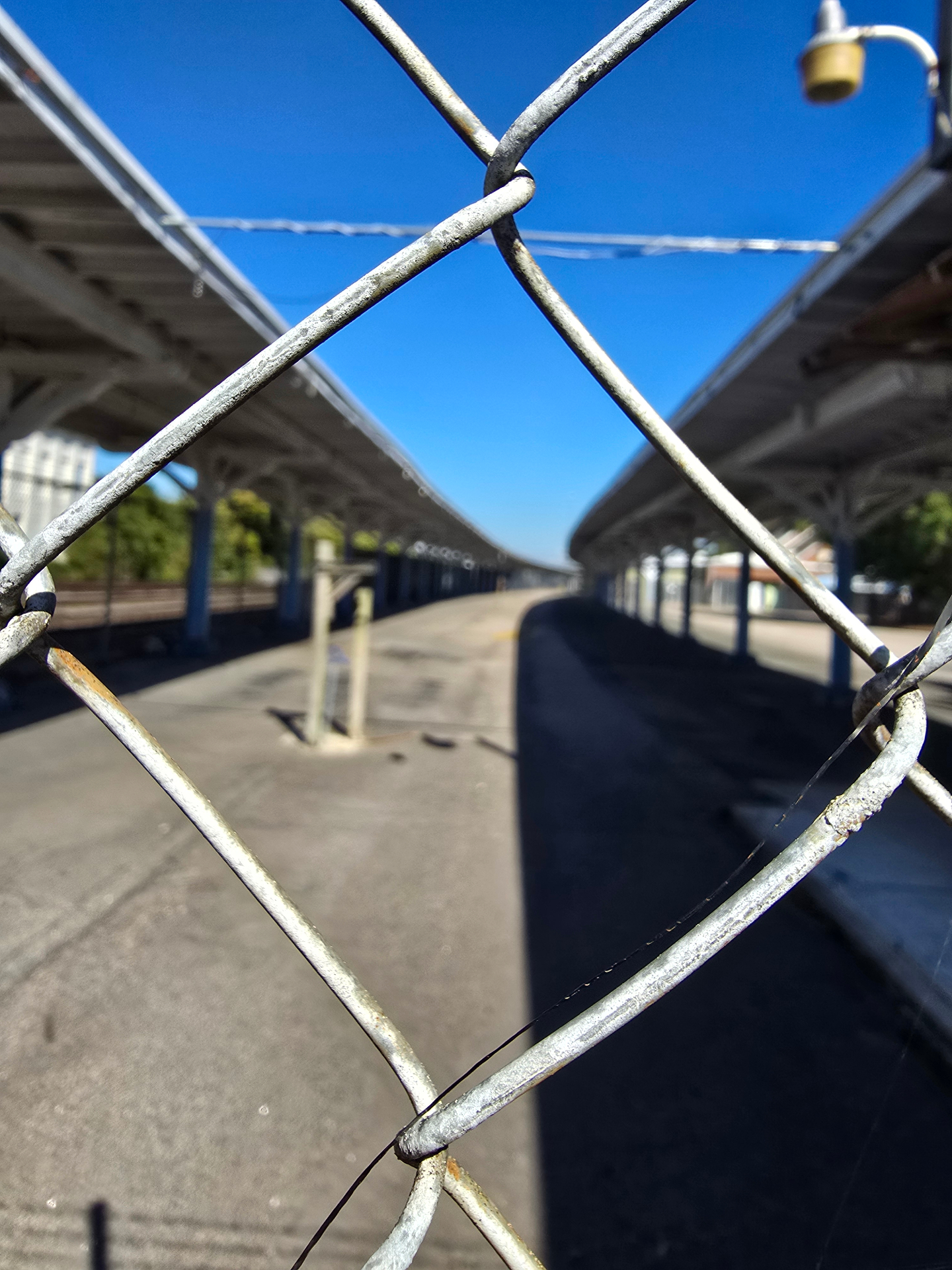 An empty sunlit train platform seen through the diamond gaps of a weathered chain-link fence