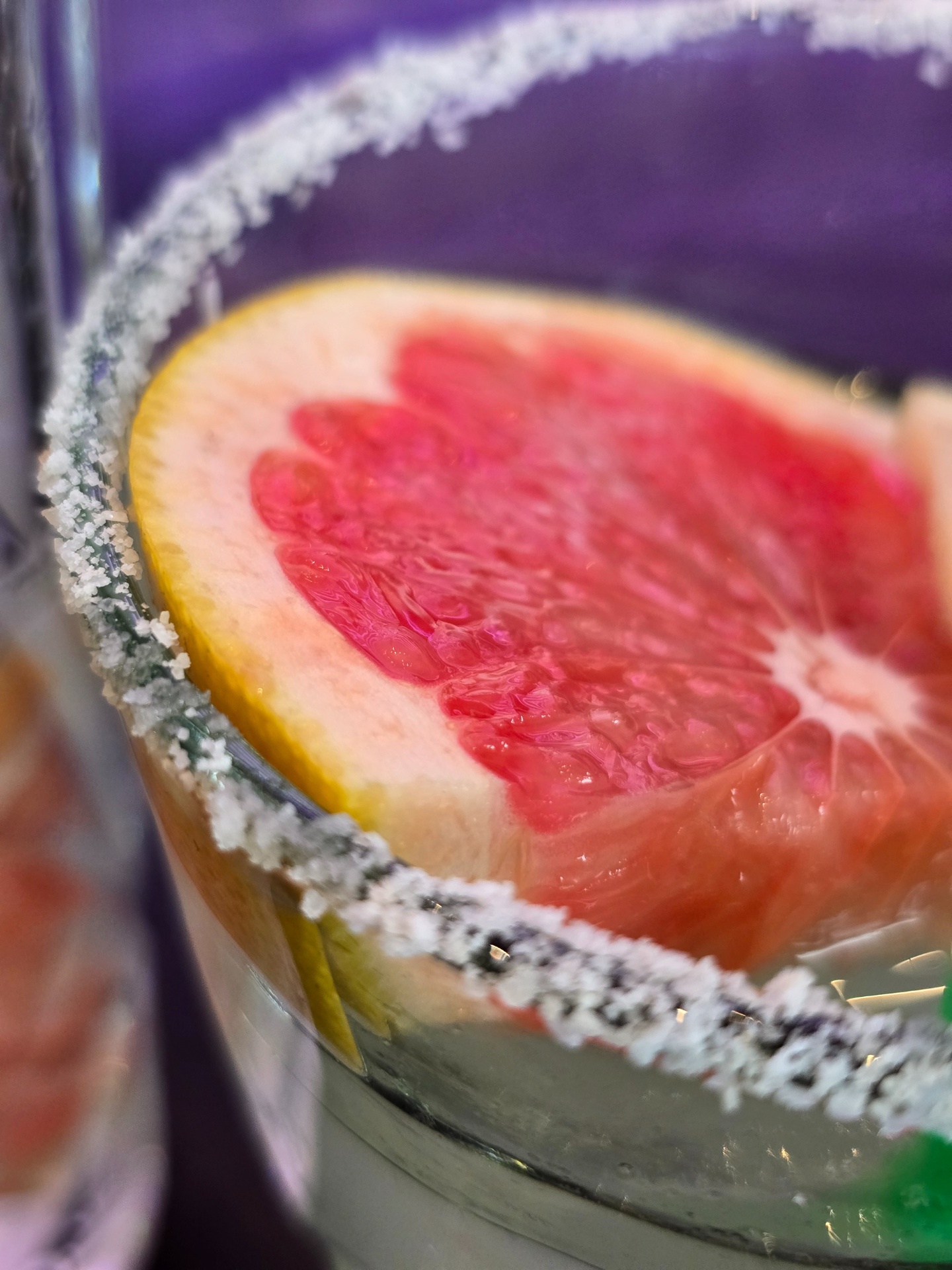 Close-up of a salt-rimmed cocktail glass with a grapefruit slice against a violet backdrop