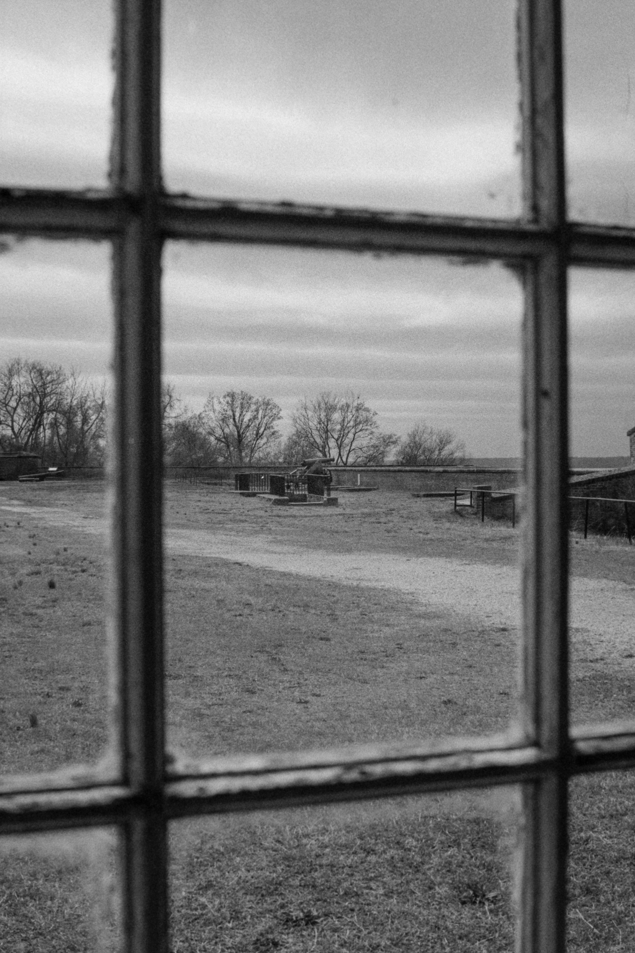 View through a paned window of a historic fort grounds with a cannon and bare trees in black and white