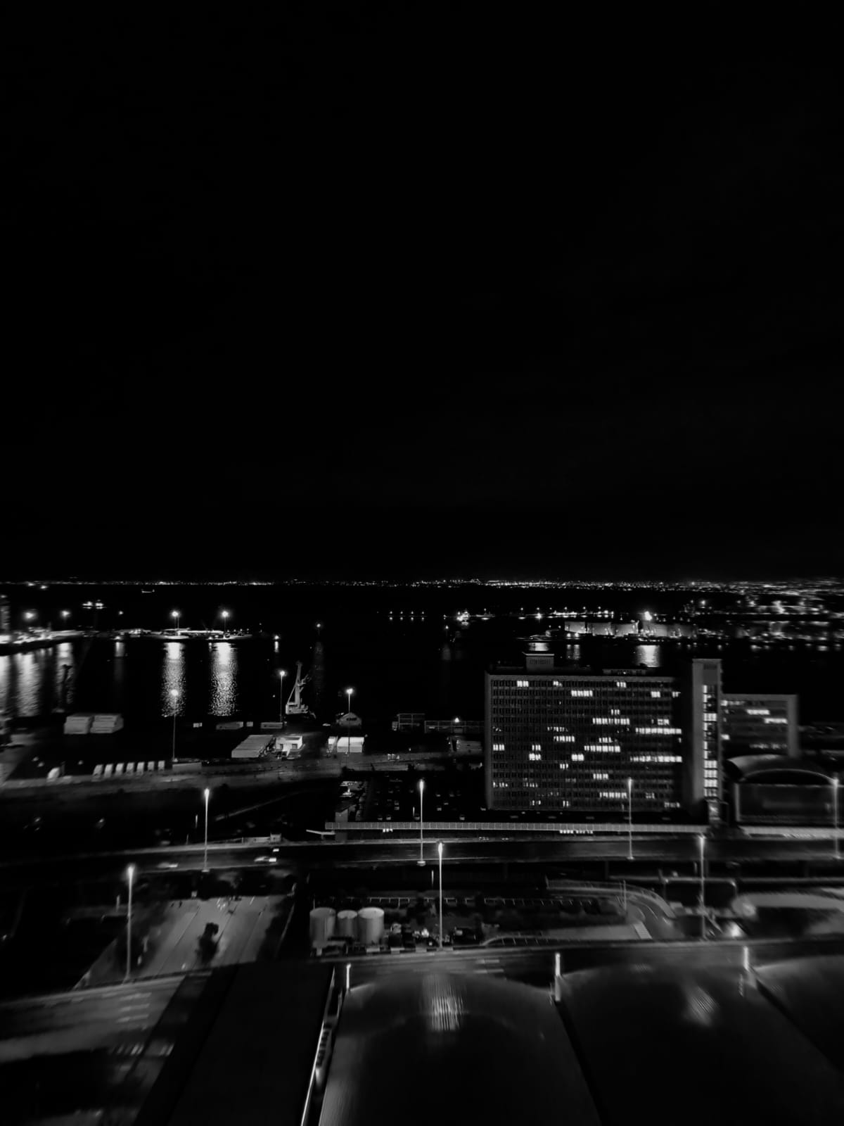 Black and white aerial night view of a harbor city with lit buildings, streetlights, and dark sky
